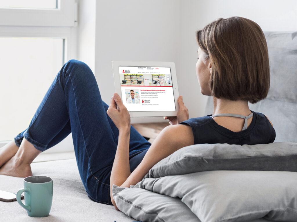 Eine junge Frau mit kurzen Haaren sitzt mit dem Rücken zur Kamera auf einem Sofa. Sie hält ein Tablet, auf dem eine Apotheken-Homepage zu sehen ist